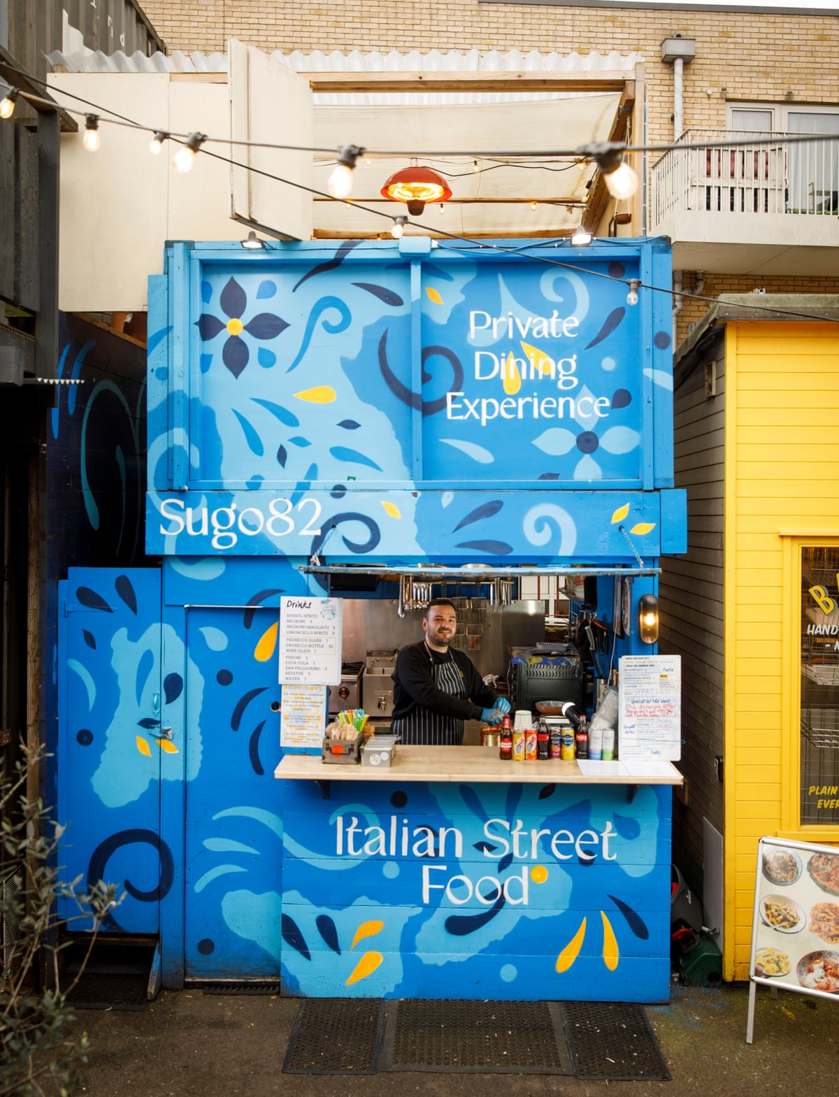 A man stands behind Sugo82's takeaway counter, which is a hatch in a blue-painted wood-panelled structure