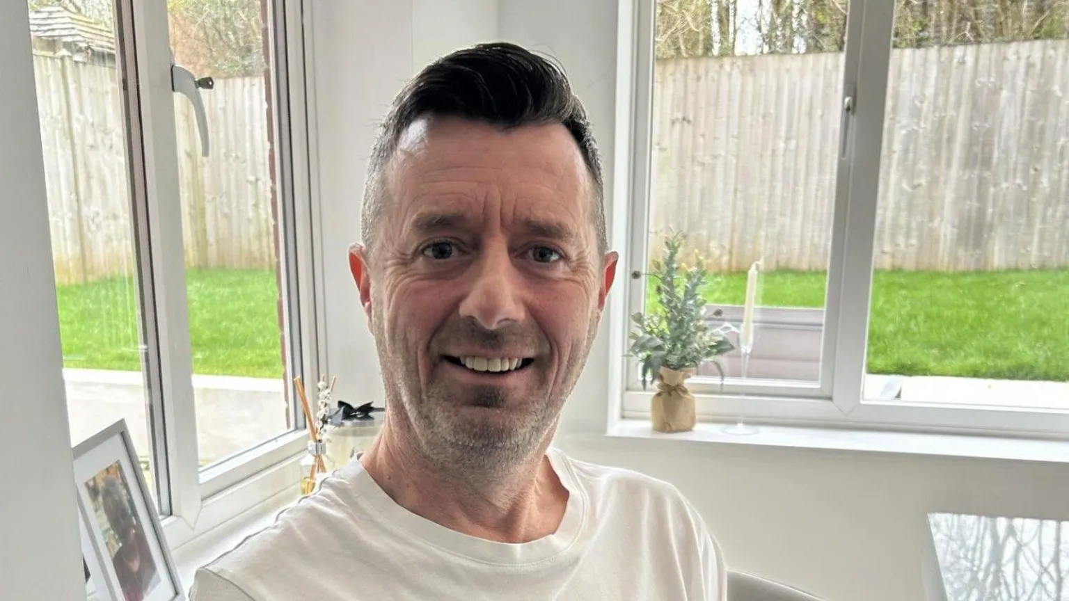 Jason Bowen sits on a chair at his dining room table, wearing a plain white t-shirt. He has short brown hair and brown eyes