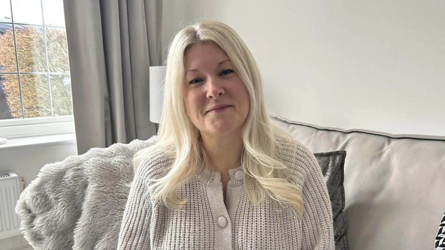 A white, middle aged woman smiles at the camera. She's sat on a cream sofa in a living room, with a window behind her. She has long blonde hair and is wearing a thick beige cardigan