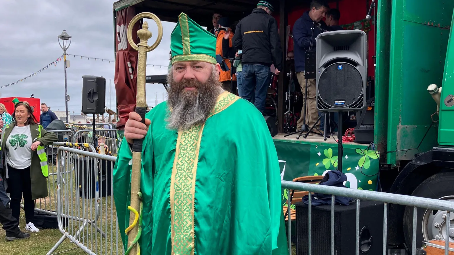 A man dressed as St Patrick. He is wearing green and gold robes and hat, holding a gold staph. 