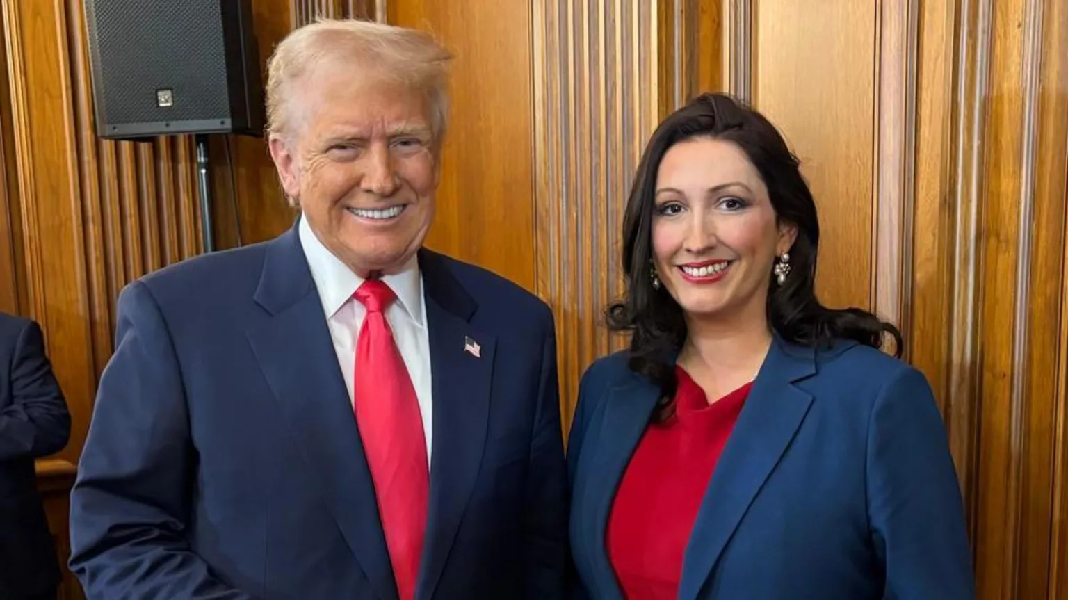 NI Executive/ Donald Trump meets Deputy First Minister Emma Little-Pengelly. She is wearing a red dress with a blue blazer. He is wearing a navy suit with a red tie. The pair are both smiling while standing in a wooden panelled room.