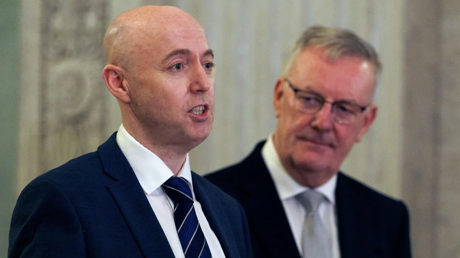 Burrows, bald and wearing a navy suit, has his lips separated but teeth clenched as he speaks. Behind him, slightly out of focus, Mike Nesbitt, wearing rectangular glasses, looks on, listening.