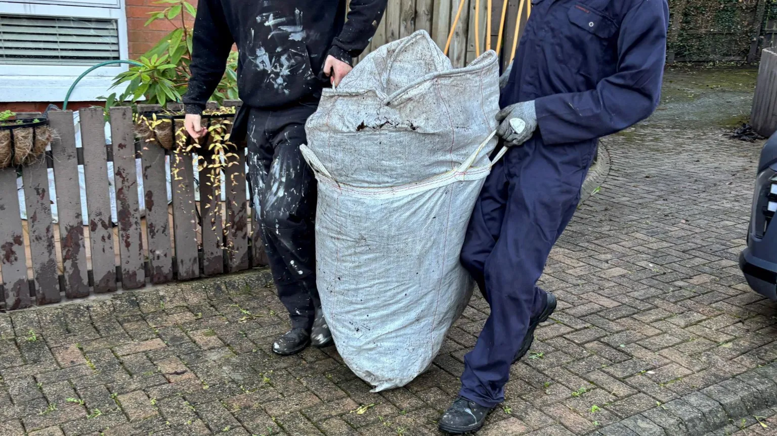 A man in a navy boiler suit, wearing black boots, hold a large white sack with both his hands, which are protected by grey gloves. He stands to the right of the bag. To the left of the bag is a man in a black hoodie, black trousers and boots. They walk in front of a dark-grey fence, on a grey brick driveway. 