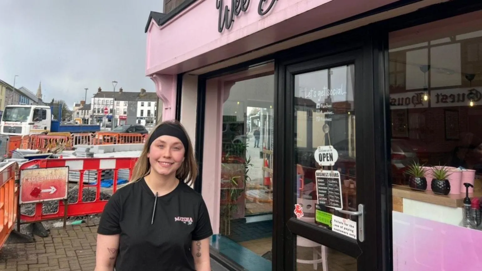 Girl with blonde hair wearing a black headband. She is standing outside of a cafe which has pink painting. Behind her is building works.