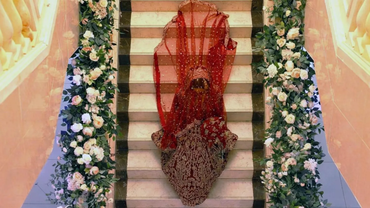 Maariya Kahn Maariya Khan, who is wearing an embellished red and gold wedding dress with a veil, is walking down the staircase which is decorated with pink flowers.
