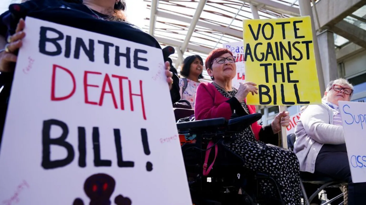Protesters outside the Scottish Parliament hold signs arguing against the assisted dying bill.