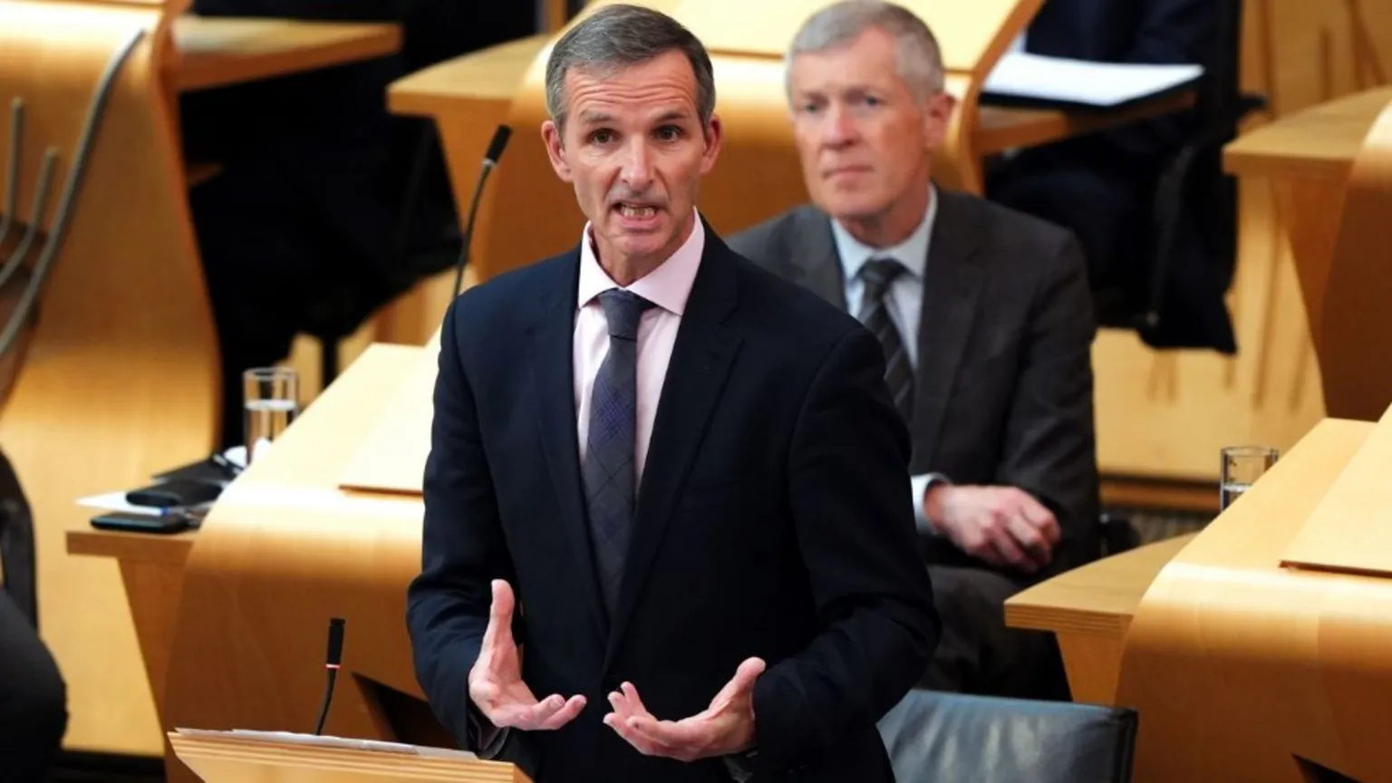 A man with greying hair speaks in the Scottish Parliament. He is wearing a dark suit, white shirt and blue tie