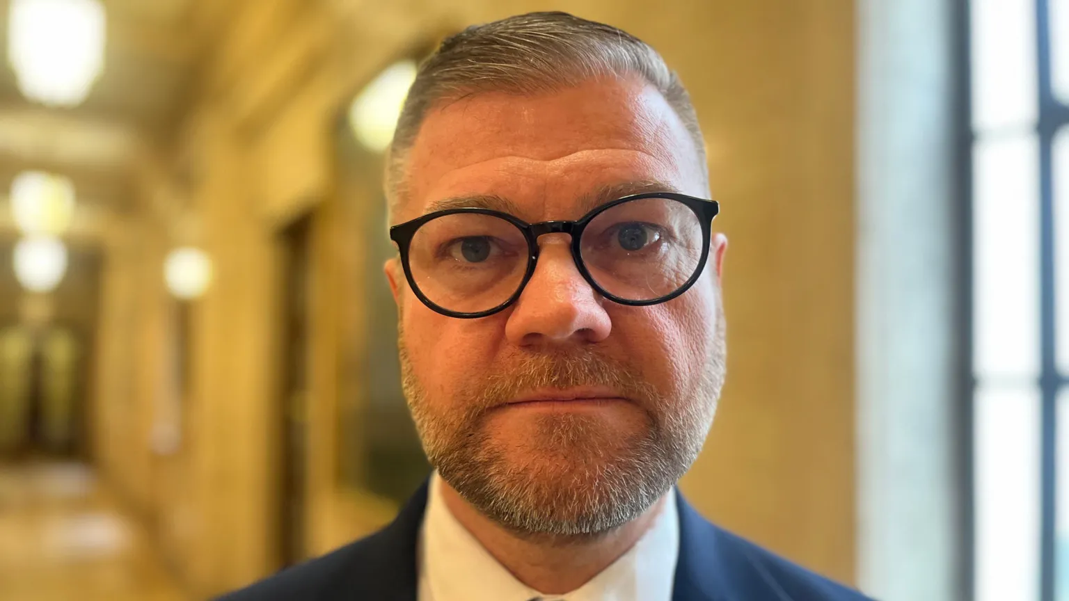 Colin looking at the camera. He has grey hair and is wearing round black framed glasses. He is wearing a white shirt and navy blazer.