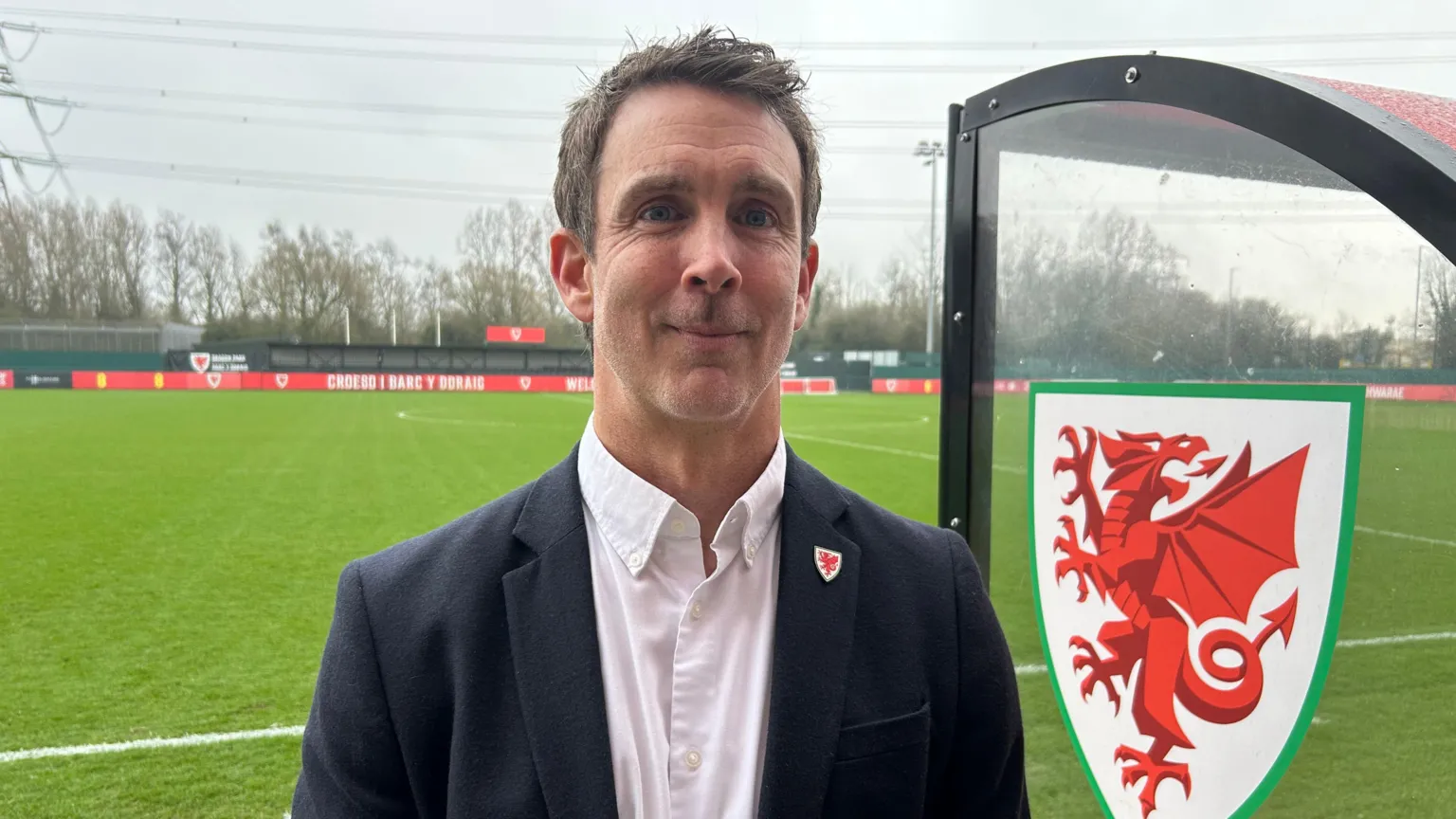 Ben Field looks at the camera. He has short dark hair and wears a white shirt and black blazer, with a pin on the lapel with a red dragon on it. An empty football pitch can be seen behind him, with a transparent shelter next to him. The shelter has a white shield emblazoned on it with a red dragon on it.