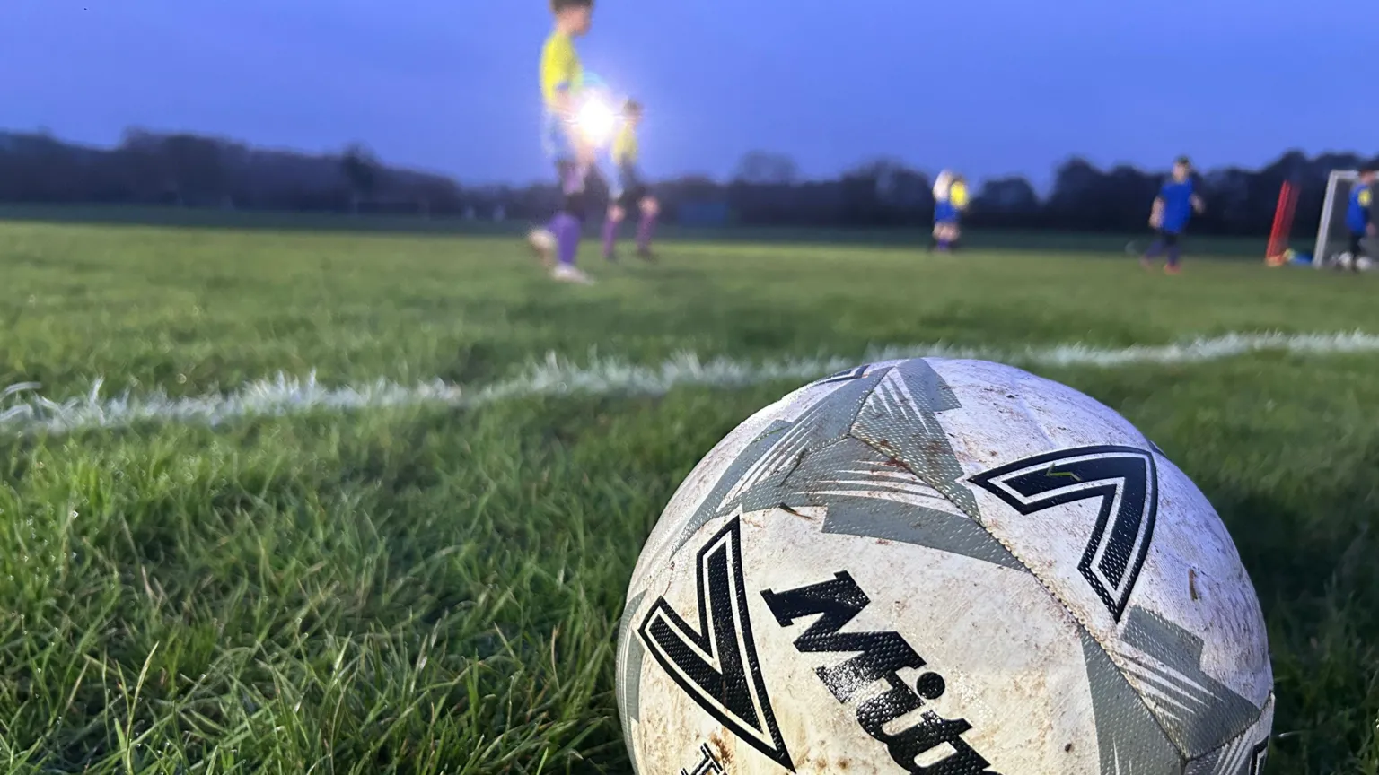 A white football on field during training sessions at Raglan Juniors FC. Players can be seen on the pitch in the background, wearing purple football socks.