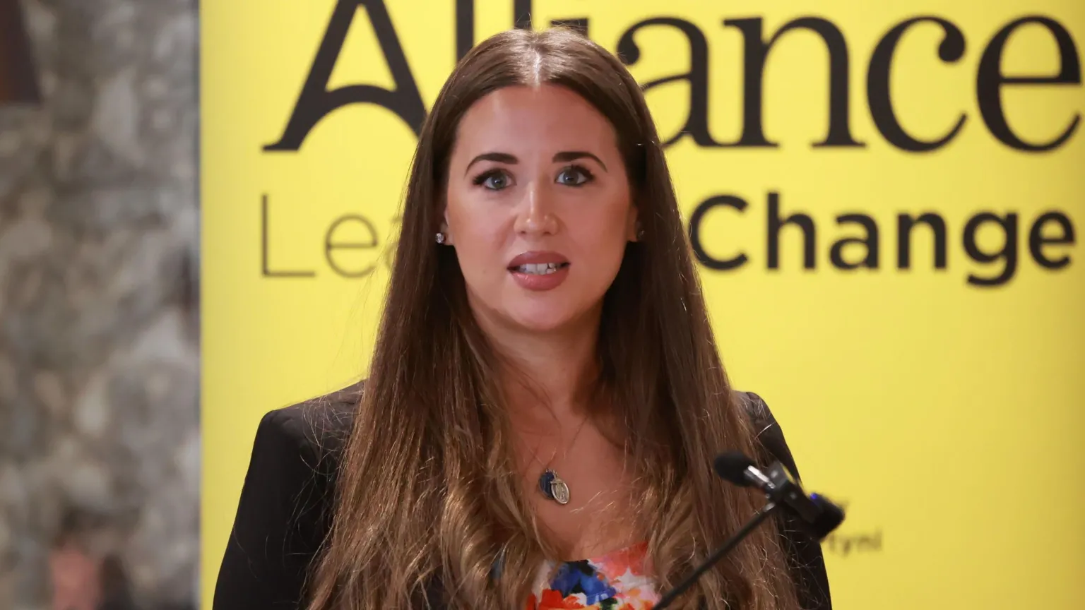 Sorcha speaking into a microphone. The background behind her is yellow. She is wearing a black blazer and multi-coloured top. She has long straight brown hair.