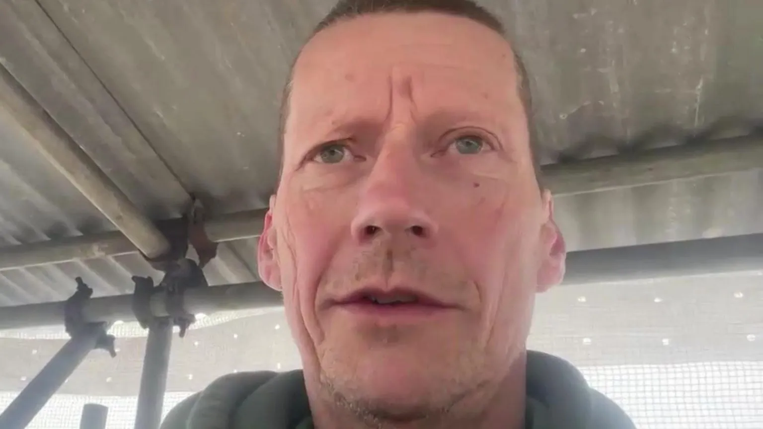 Gerald Salvat stands next to some scaffolding with corrugated iron on top of it. He is has stubble and short dark hair.