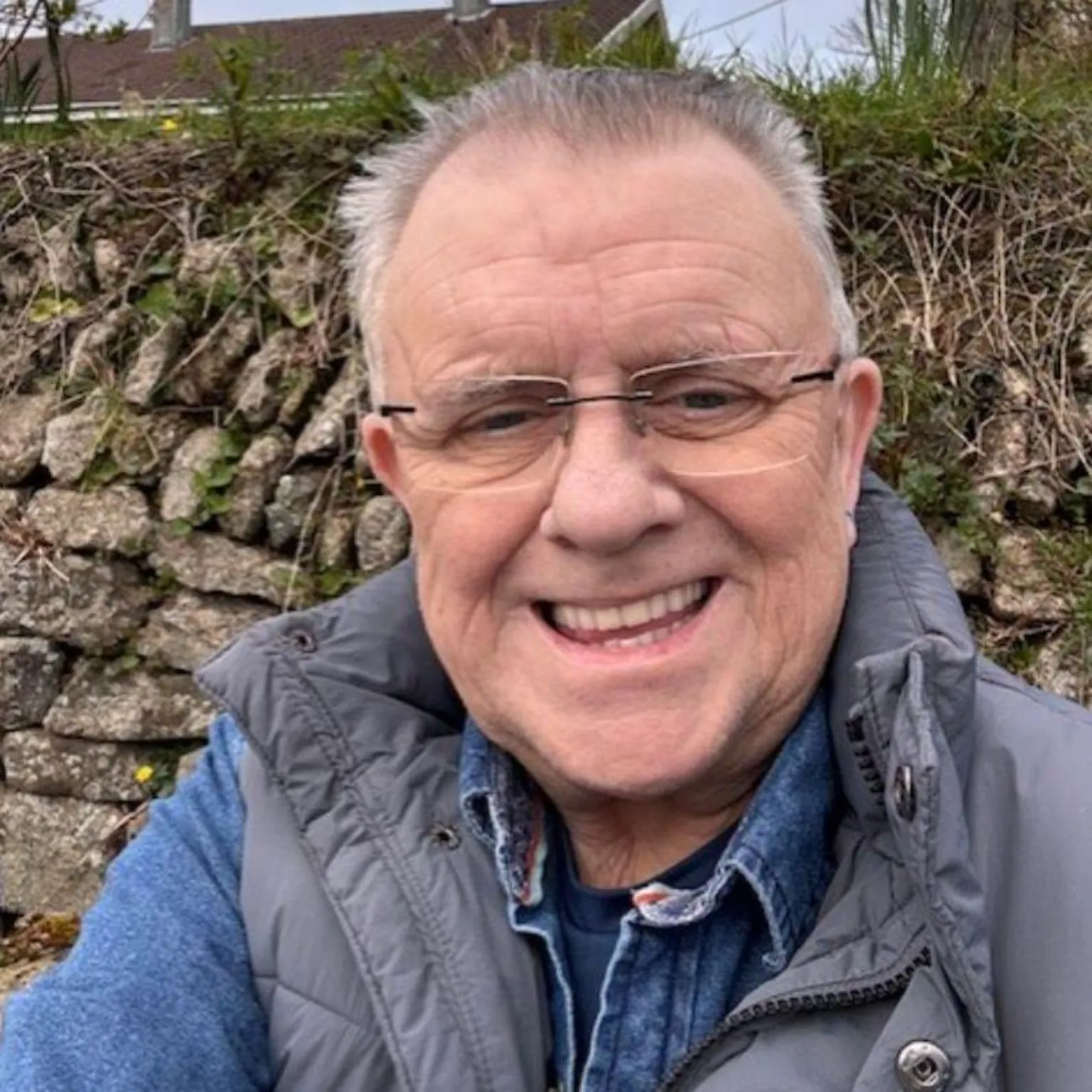 Alan Dickinson Alan Dickinson smiles at the camera while being stood in front of a hedged wall which has plants growing out of it. The roof of a house can be seen in the distance. Dickinson, who is 67, has glasses on along with a blue denim shirt and grey gilet.