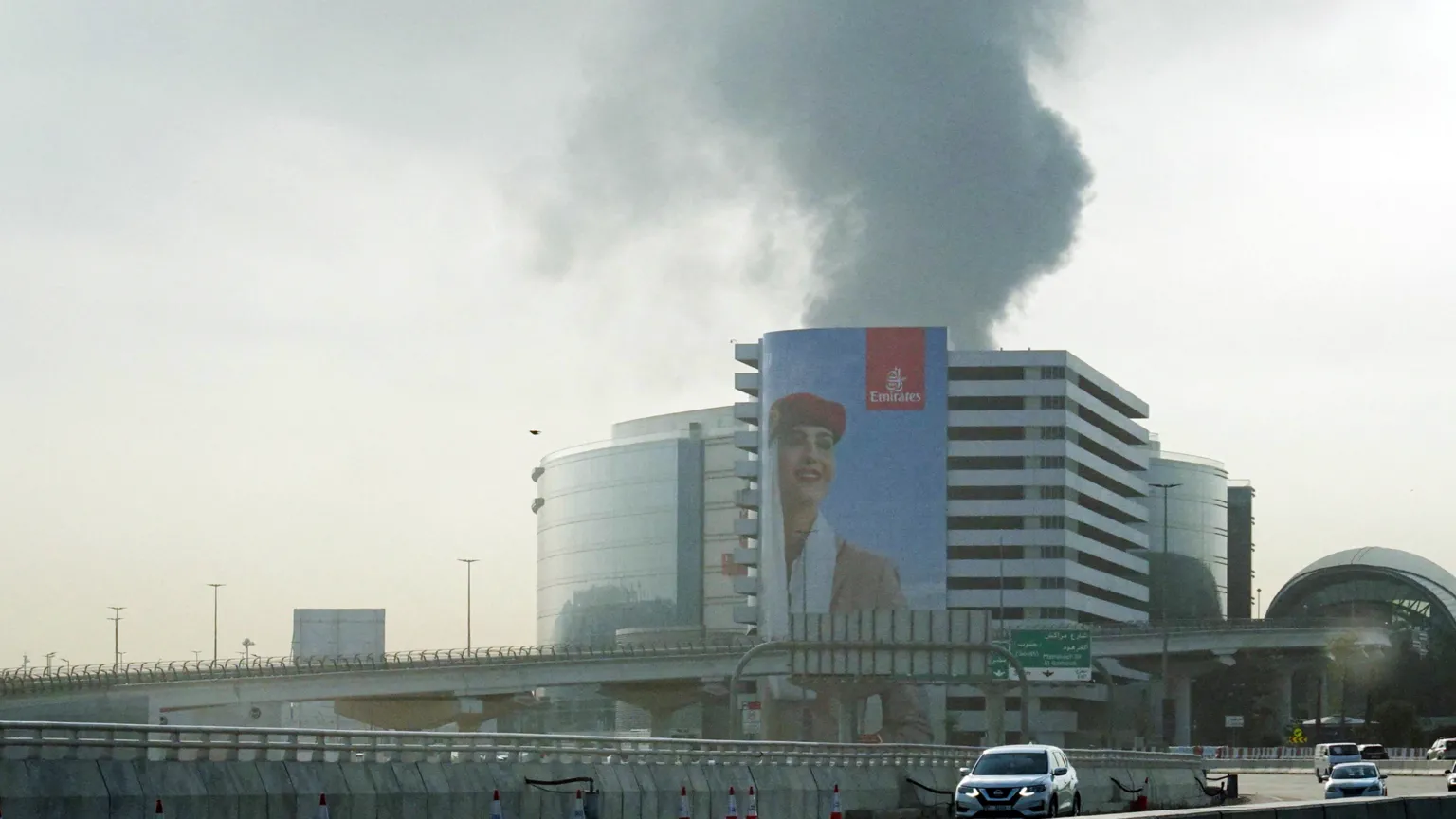 Black smoke rising from a building in Dubai. There is a huge Emirates advert on the side of the building featuring a female member of cabin crew. There are roads and cars in front of the building.