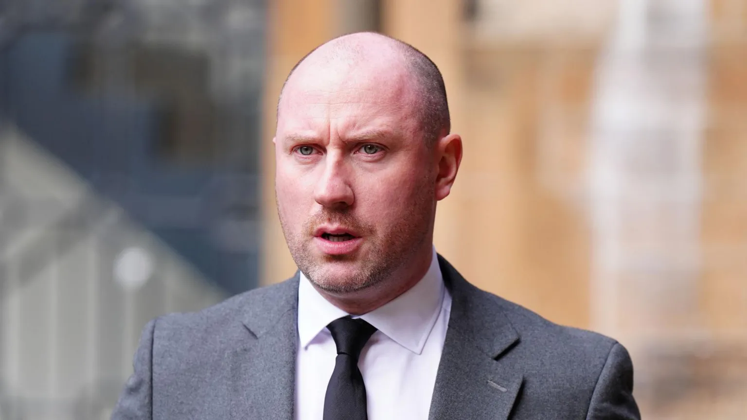  Neil Gray is bald and wears a grey suit with a white shirt and black tie. He looks ahead while walking outside a building
