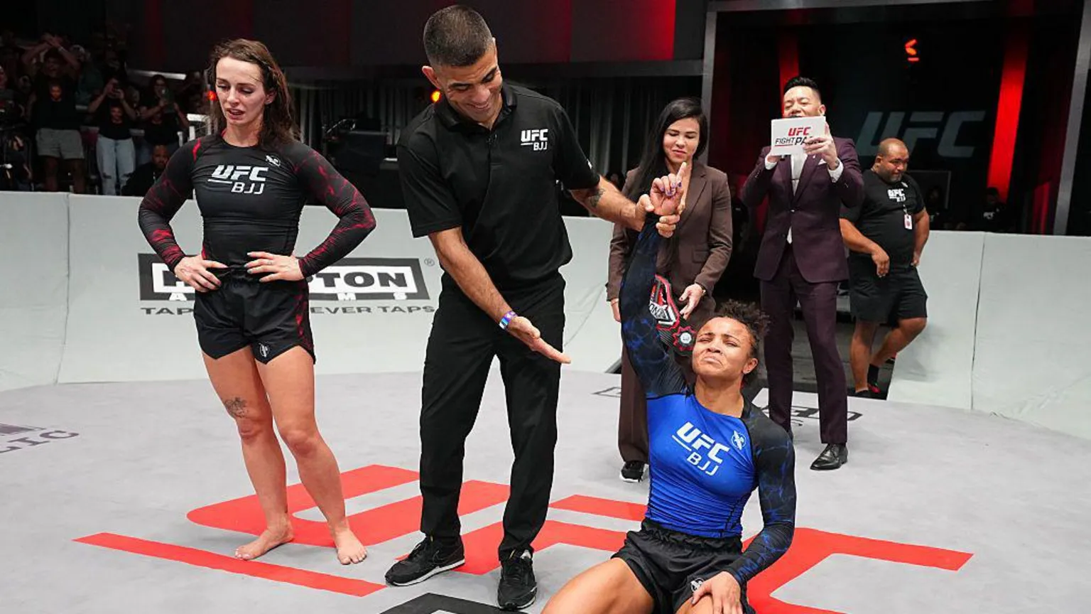  Ffion Davies on the left looks at the floor with her hands on her hips. On the right, Cassia Moura has her hand raised by the referee with an expression of relief on her face after her victory in the women's bantamweight championship match