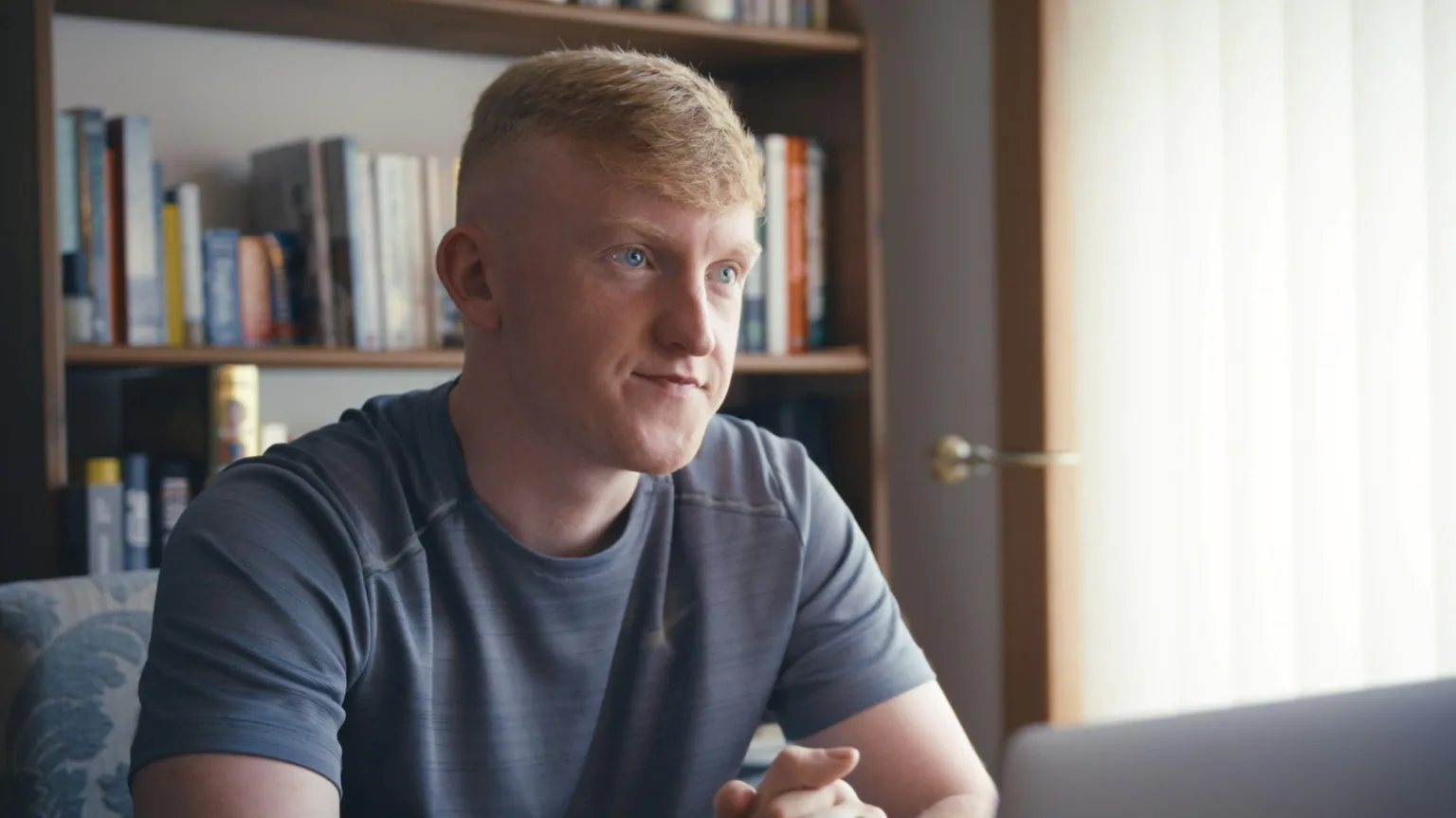 Head and shoulders image of Blair McNally. He short red hair and blue eyes and is wearing a grey, short-sleeved sports top. He's sitting in front of a bookcase.