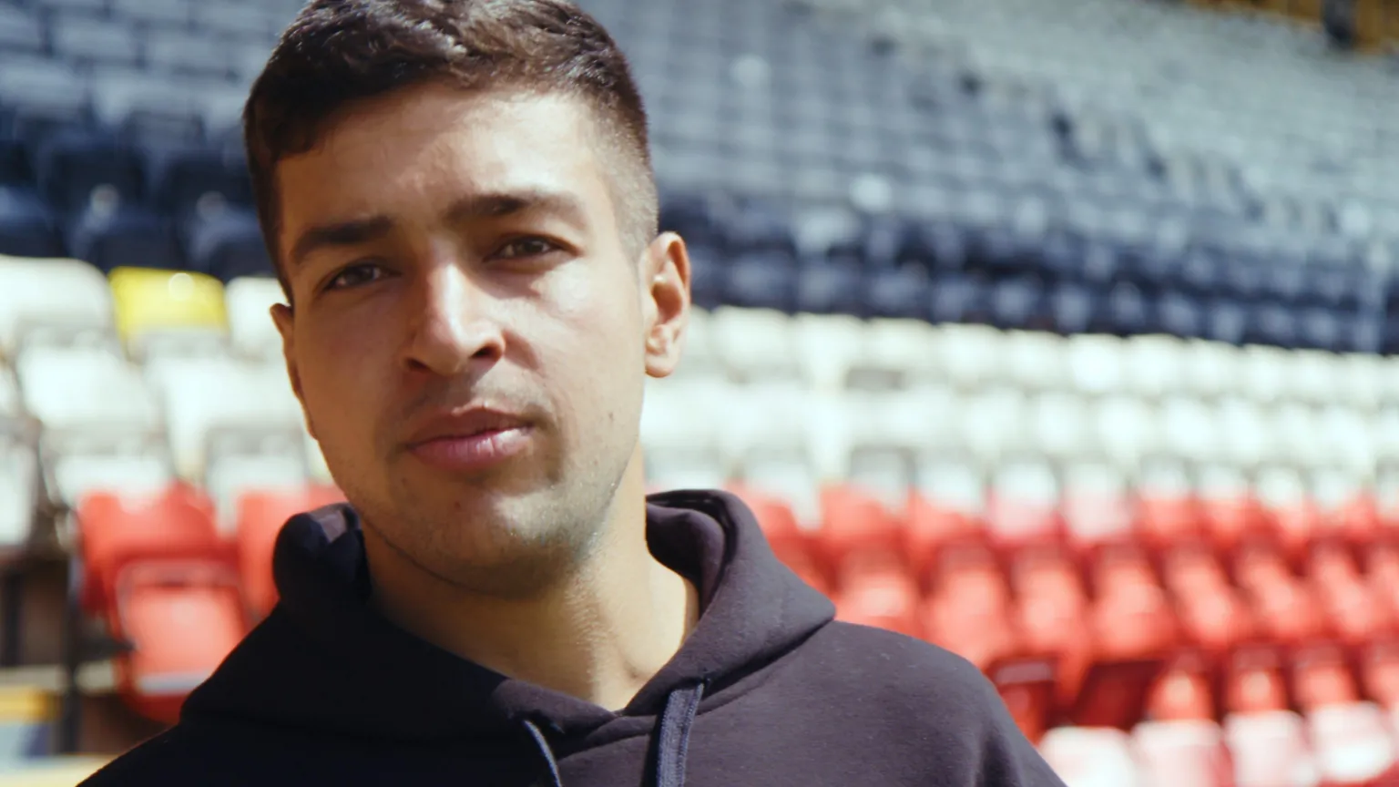 Head and shoulders image of Manpreet Singh. He has short dark hair and is wearing a navy hoodie. He's standing in front of black, red and white seats in a football stadium.