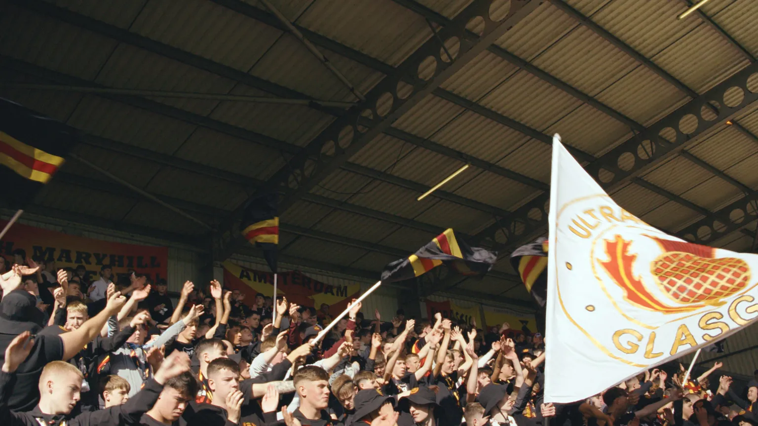 Patrick Thistle Ultras in a stand at the Firhill Stadium waving flags balck, red and yellow flags and singing with their hands in the air. There is a larger white, red and yellow flag on the right side of the crowd.