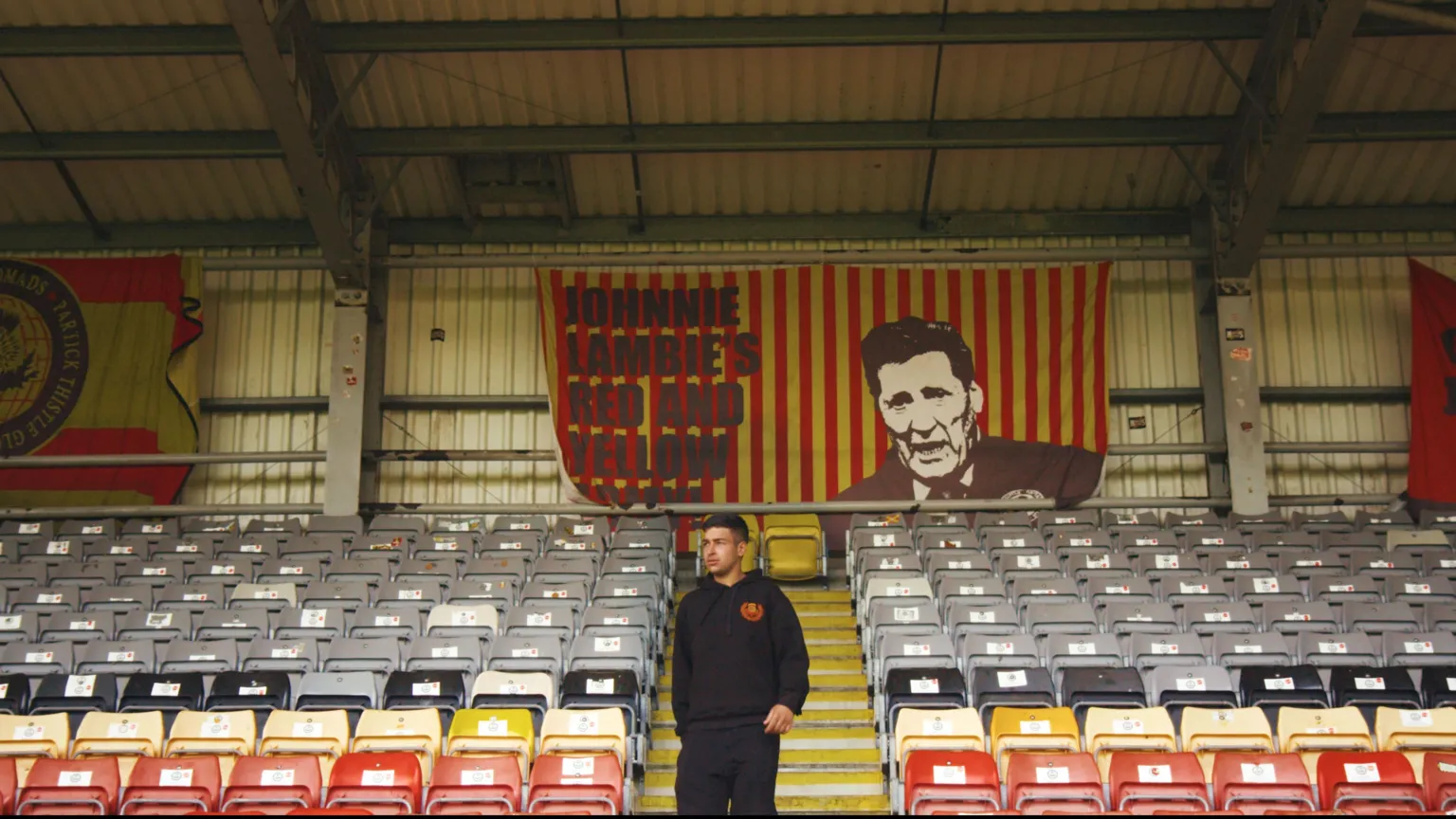 Image of Manpreet Singh at Firhill Stadium. He's standing in the middle of one of the stands with rows of empty grey, black, yellow and read seats to either side of him. There are three banners on the back wall. One reads