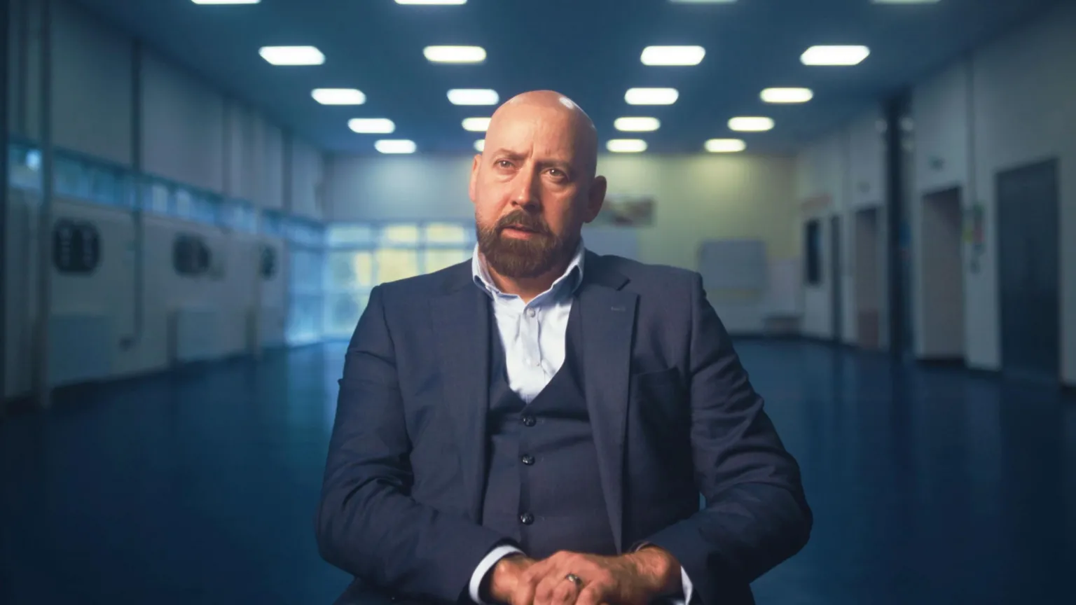 Image of David Kennedy. He is sitting in a brightly lit room staring straight at the camera with his hands in his lap. He is bald, with a dark beard and moustache and is wearing a three-piece navy suit and pale blue shirt.