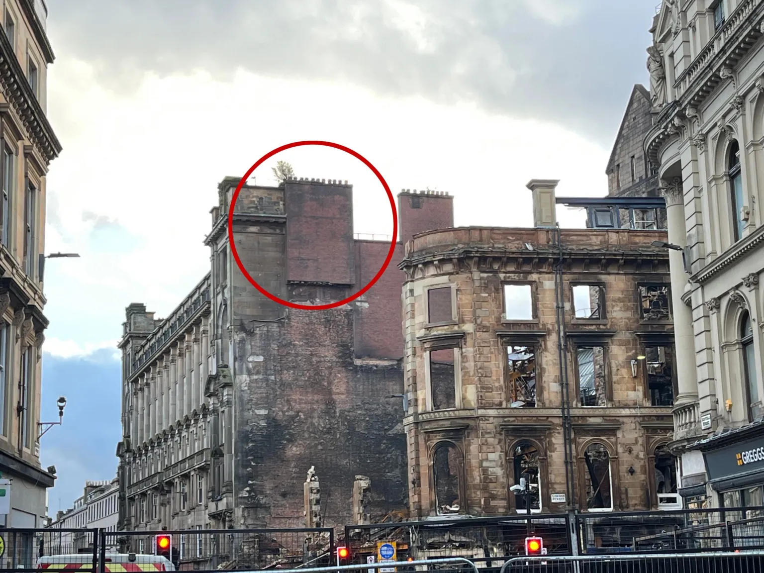 The fire-ravaged shell of a building on the corner of a street in the centre of Glasgow. There is a chimney at the top of the building, which was previously attached to the building which has now collapsed. It appears to hang in mid-air and has a red circle around it.