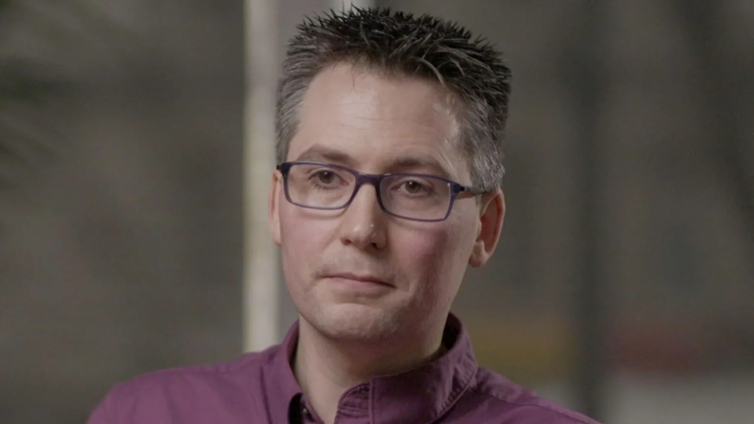 Chris Wynne has dark hair, which is grey at the sides, is wearing a purple shirt and purple, rectangle framed glasses. He is looking to the side of the camera. The background of the image is blurred.