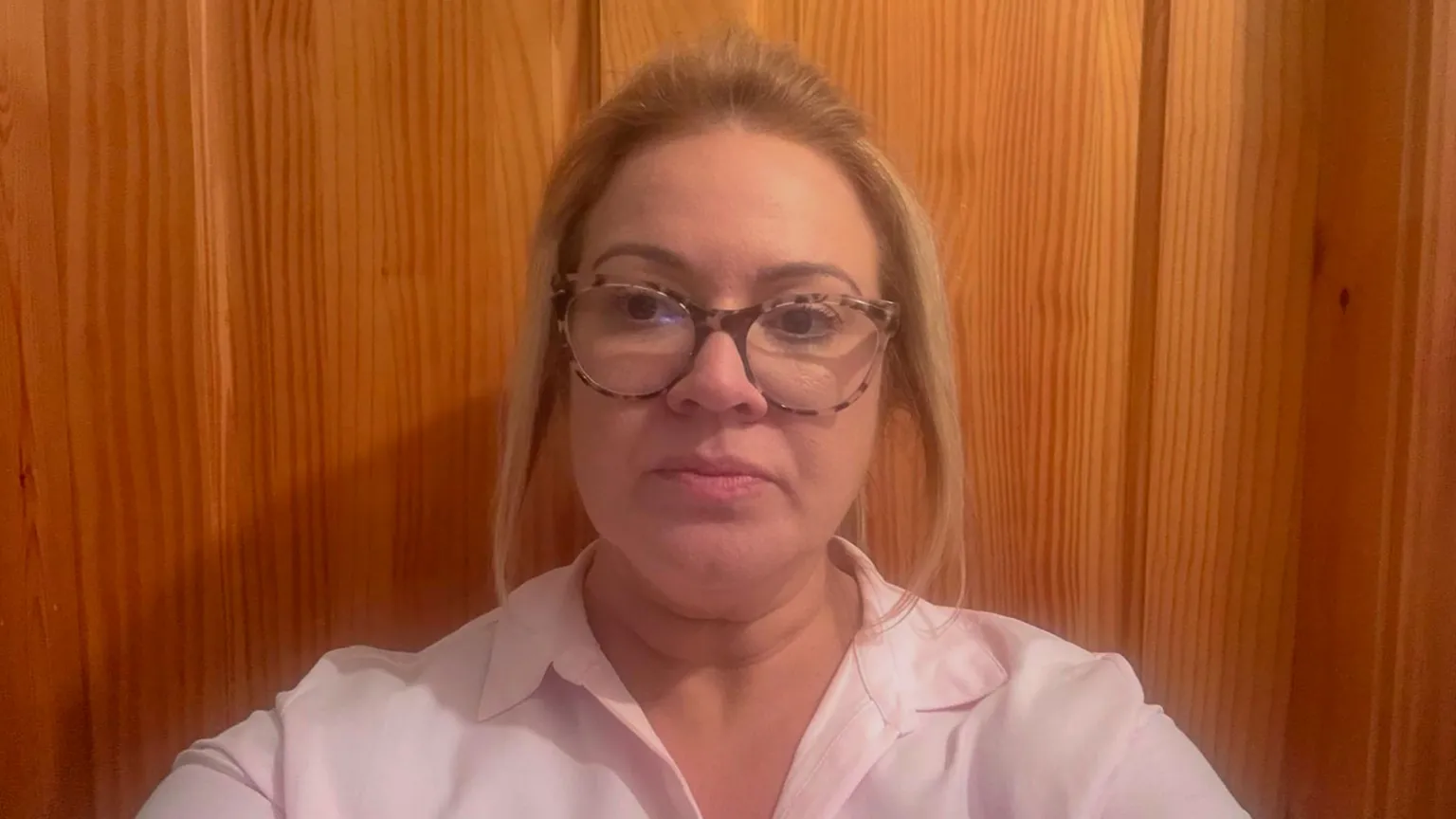 A woman with mid length blonde hair, wearing a pink open collar shirt, is looking directly at the camera. She is standing in front of a pine coloured wooden door and is wearing large, round framed glasses with a brown leopard print on the rim.