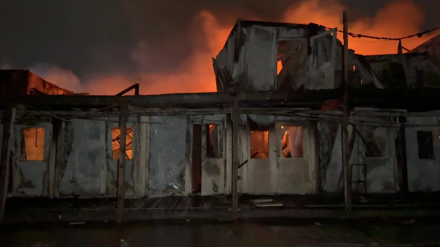 A building on fire at night. It has been badly damaged, with flames and smoke glowing orange behind the shell of the building.