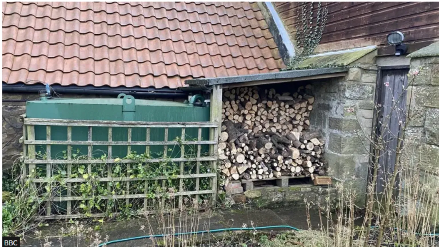 A heating oil tank sits next to a pile of wood in the corner of an old shed. A wooden door is just to the right of the woodpile 