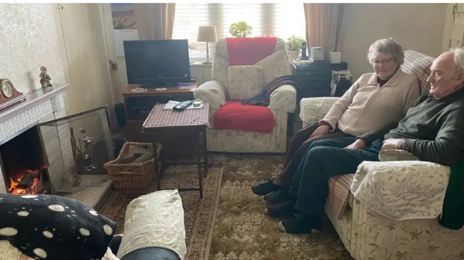An elderly couple sit in front of the fire in an old-fashioned sitting room. The room is small but cosy with lots of rugs and a wood basket and fireguard 