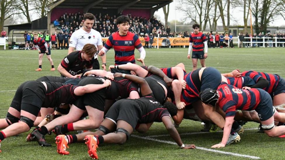 Rugby players from NSB and Campion School are in a scrum and the ball is about to be fed. A referee in a white shirt is standing next to the scrum and there is a crowd of people watching in the background.