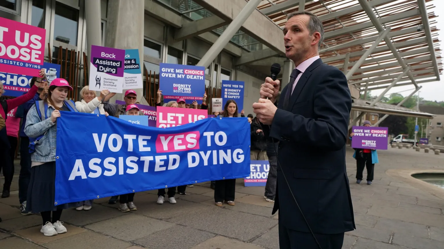  McArthur standing outside the Scottish Parliament speaking into a microphone. Behind him are protesters in favour of the bill, holding a banner and placards.
