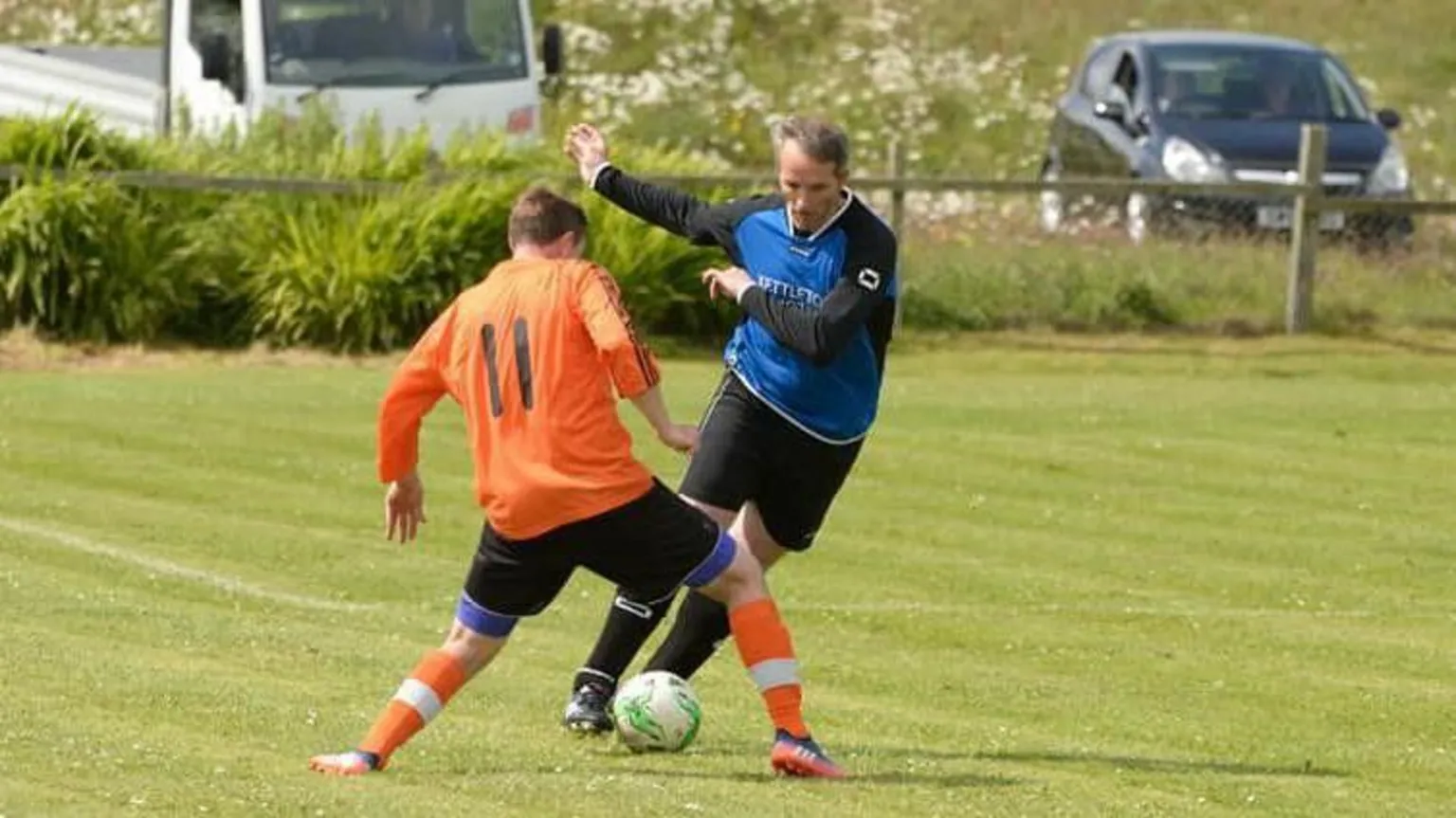 McArthur family Liam McArthur playing football on a grass pitch. He is wearing a blue and black football strip and is trying to take the ball past an opponent in an orange strip.