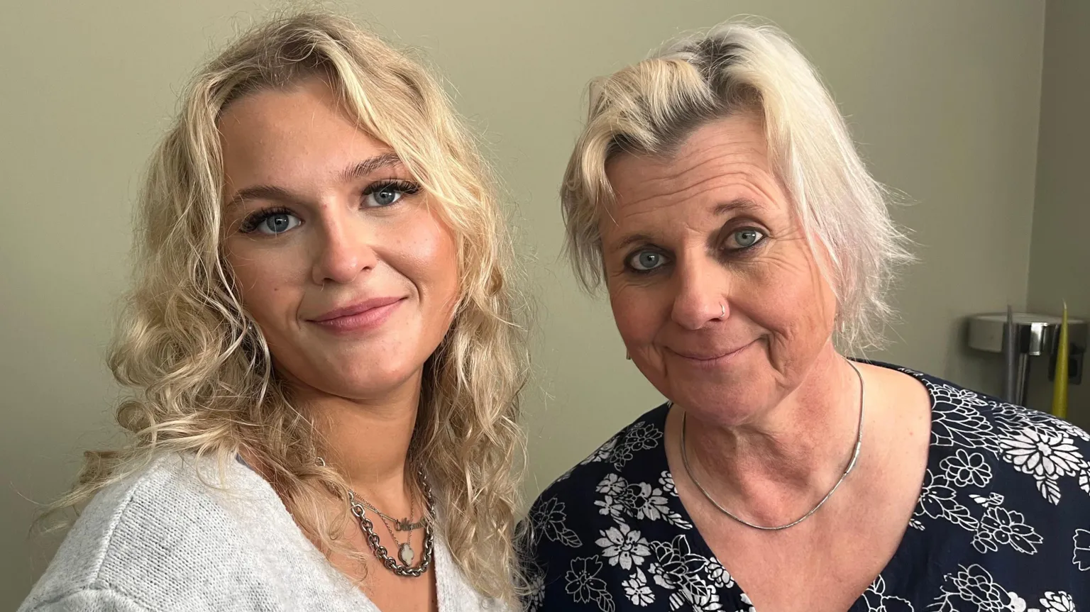 Young woman with blonde hair wearing a grey v neck jumper alongside an older woman who's her woman who also has blonde hand and is wearing a black v neck top with white flowers on. Both are smiling at the camera. 