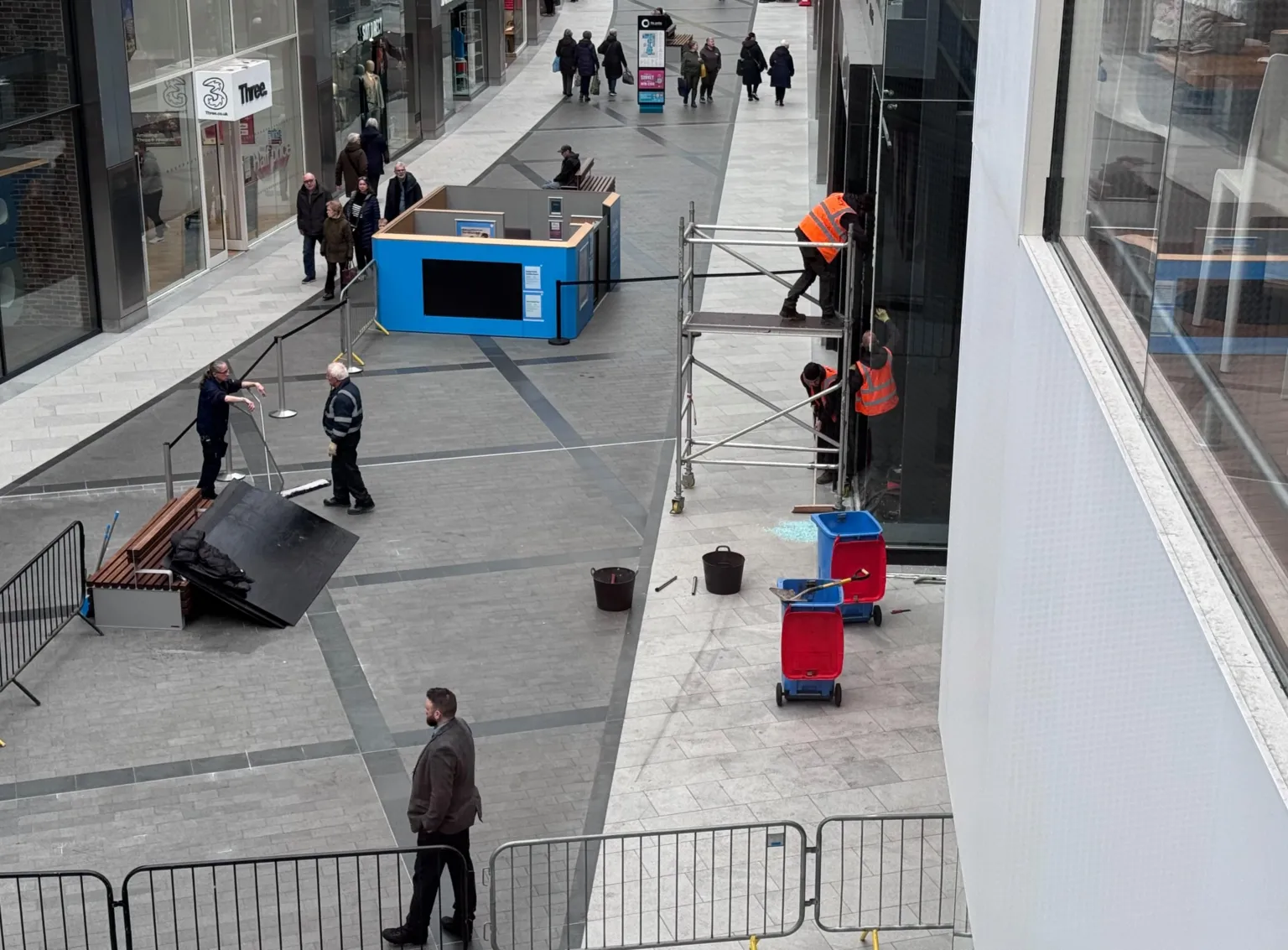 Workers in high‑visibility clothing securing damaged glass doors with scaffolding inside the Almondvale Shopping Centre in Livingston, with barriers and debris on the ground after a ram‑raid incident.