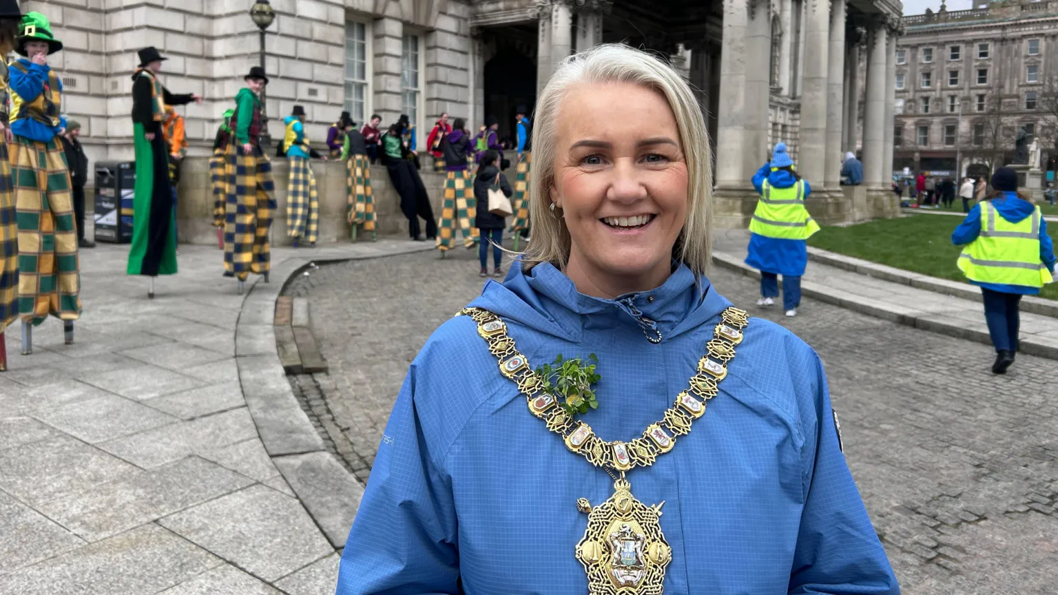 Tracy Kelly is wearing a large gold chain of office around her neck and shamrock has been pinned to her blue raincoat. There are people on stilts behind her and some people wearing high viz jackets. The city hall is in the background, with large grey pillars. 