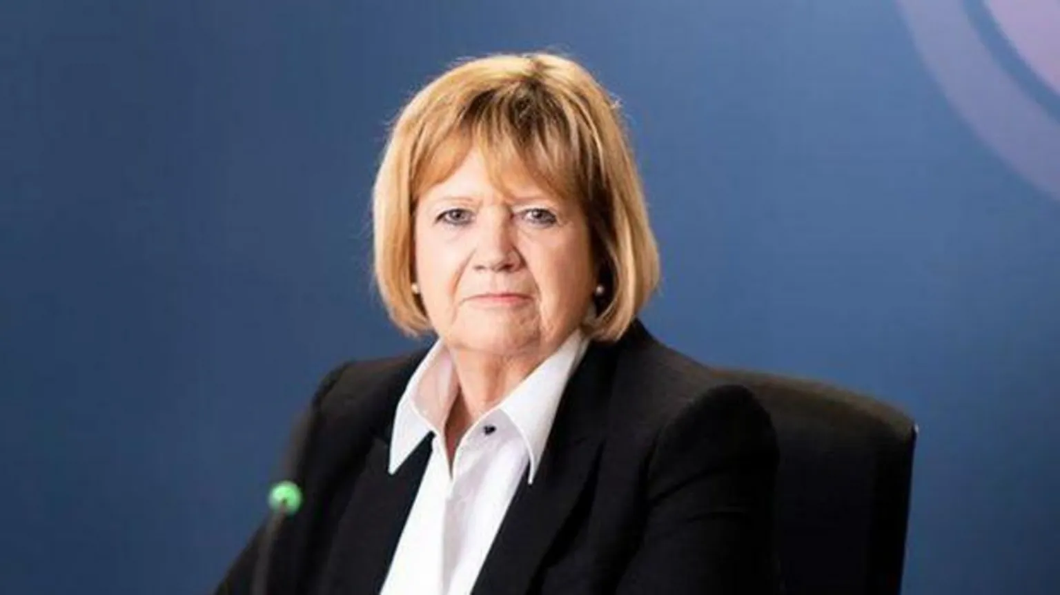 Pirahna Photography Baroness Hallett in front of a blue backdrop featuring the pink, purple and white Covid-19 inquiry logo. 