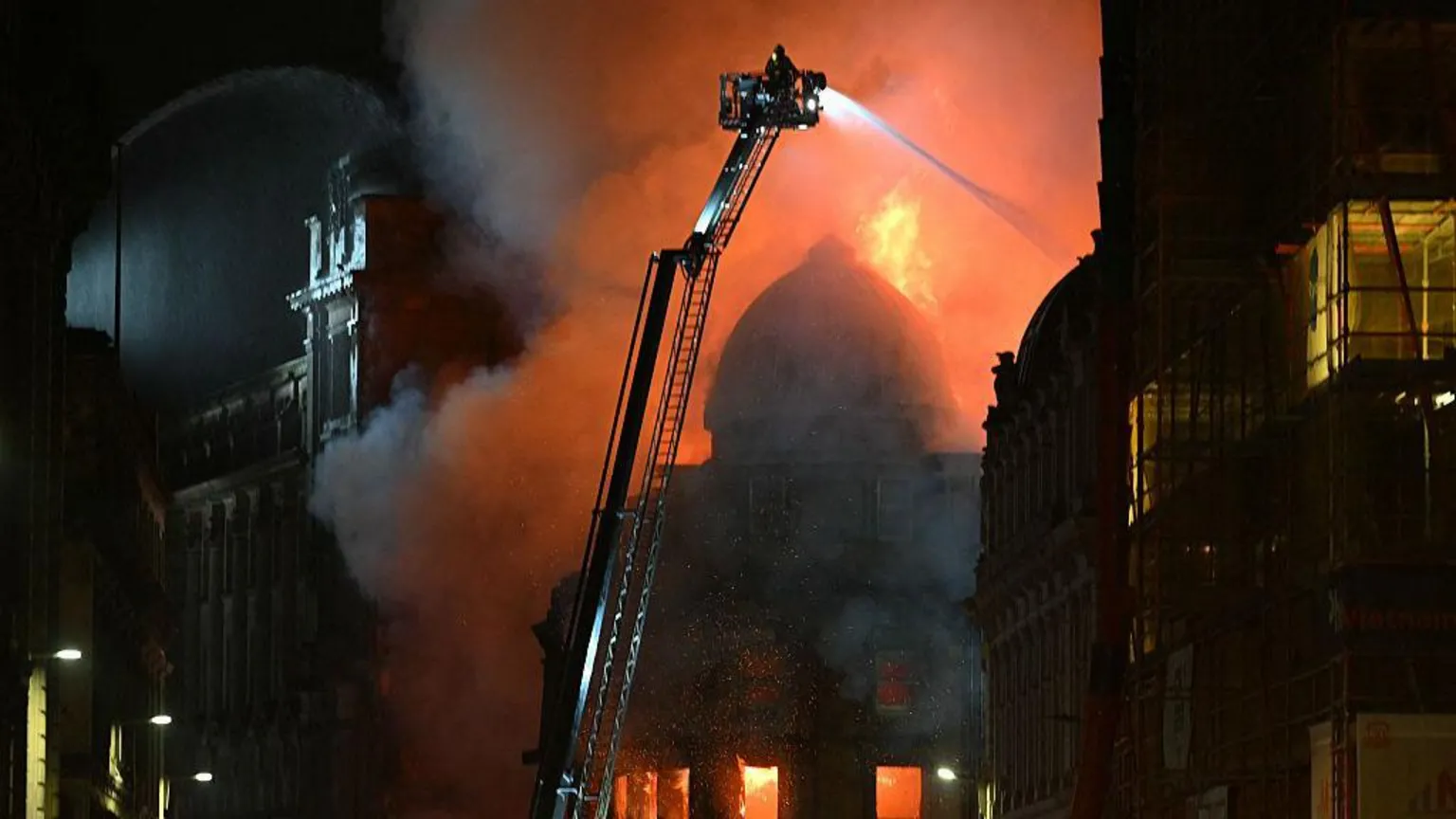  A dome of a Victorian building on fire. There is a crane with a firefighter spraying water on the flames and other buildings surrounding it in darkness.