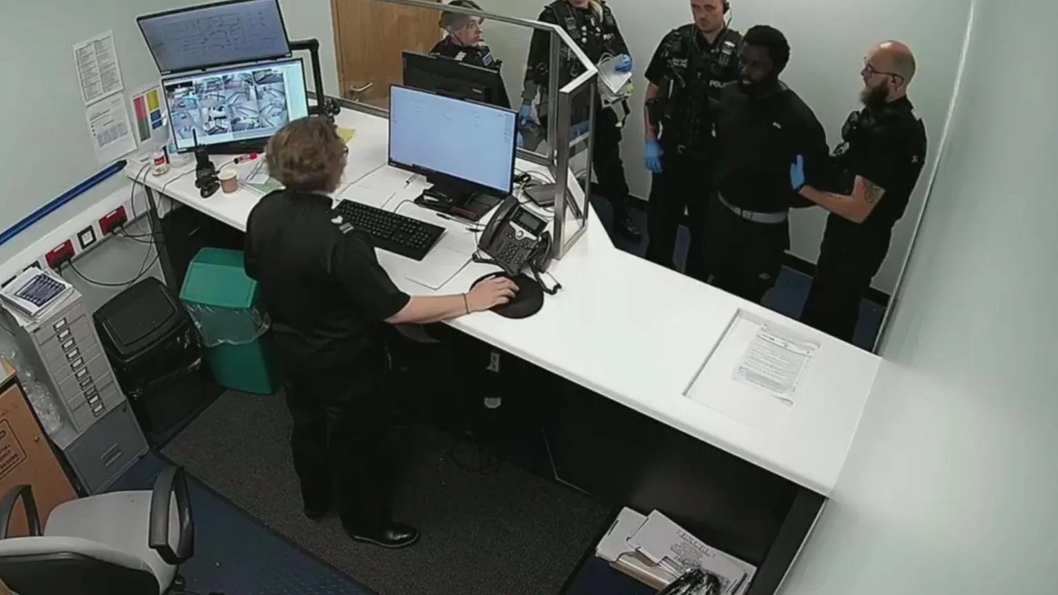 Nottinghamshire Police CCTV image of a police custody reception, with four officers and Valdo Calocane stood on the far side of the desk, and the custody sergeant nearer the camera