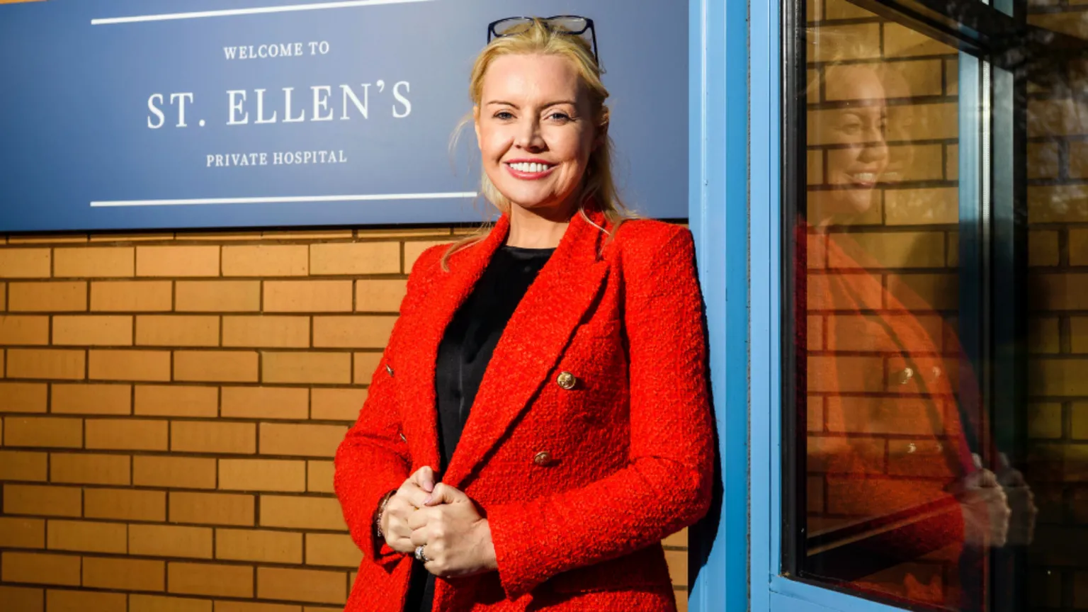 Gill Baird, who has blonde hair tied back, smiles at the camera while standing outside a brick building. She is wearing a red jacket over a black top. 