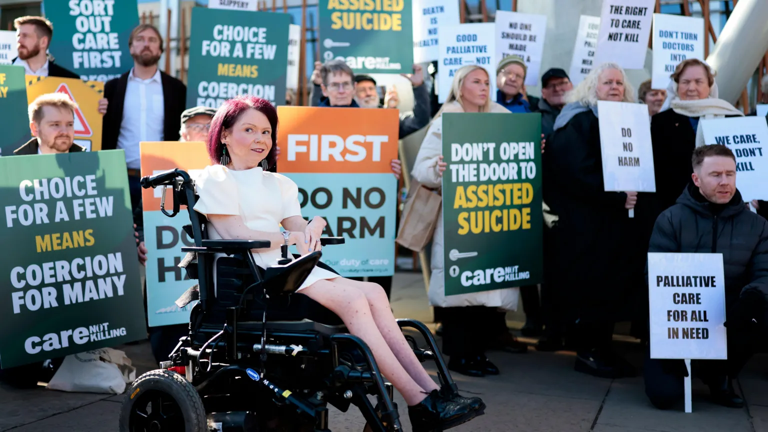  A woman in a wheelchair in front of a group of protesters holding placards arguing against assisted dying 