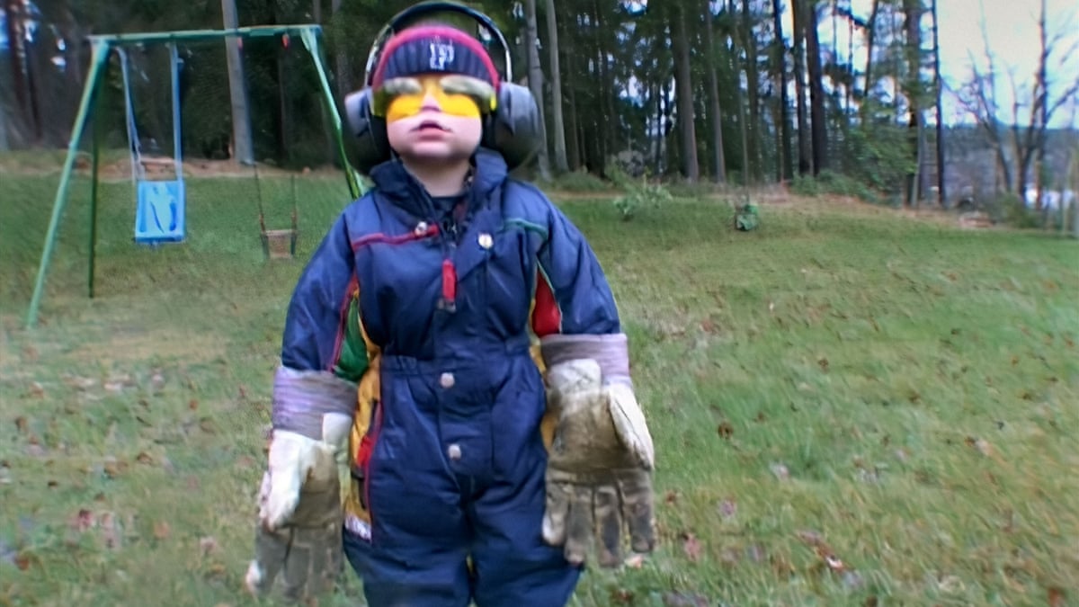 A young child in a garden wearing a blue jumpsuit, huge gardening gloves, yellow goggles and large headphones