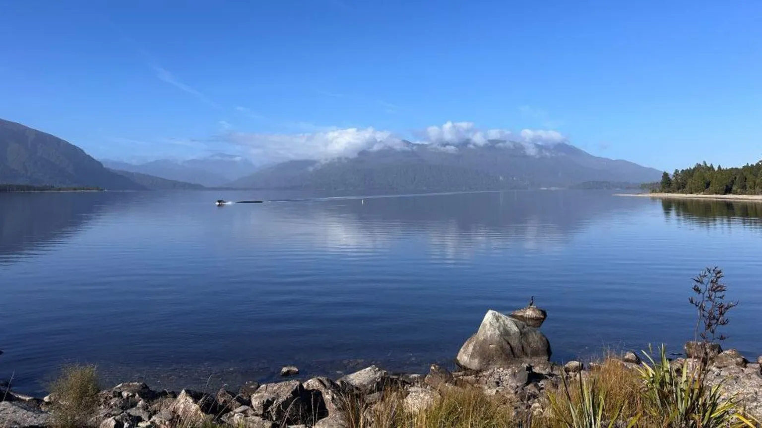Lori Carnochan A landscape: Clear water, with hills in the background. The sky is blue but there are some clouds around the top of the mountains. 