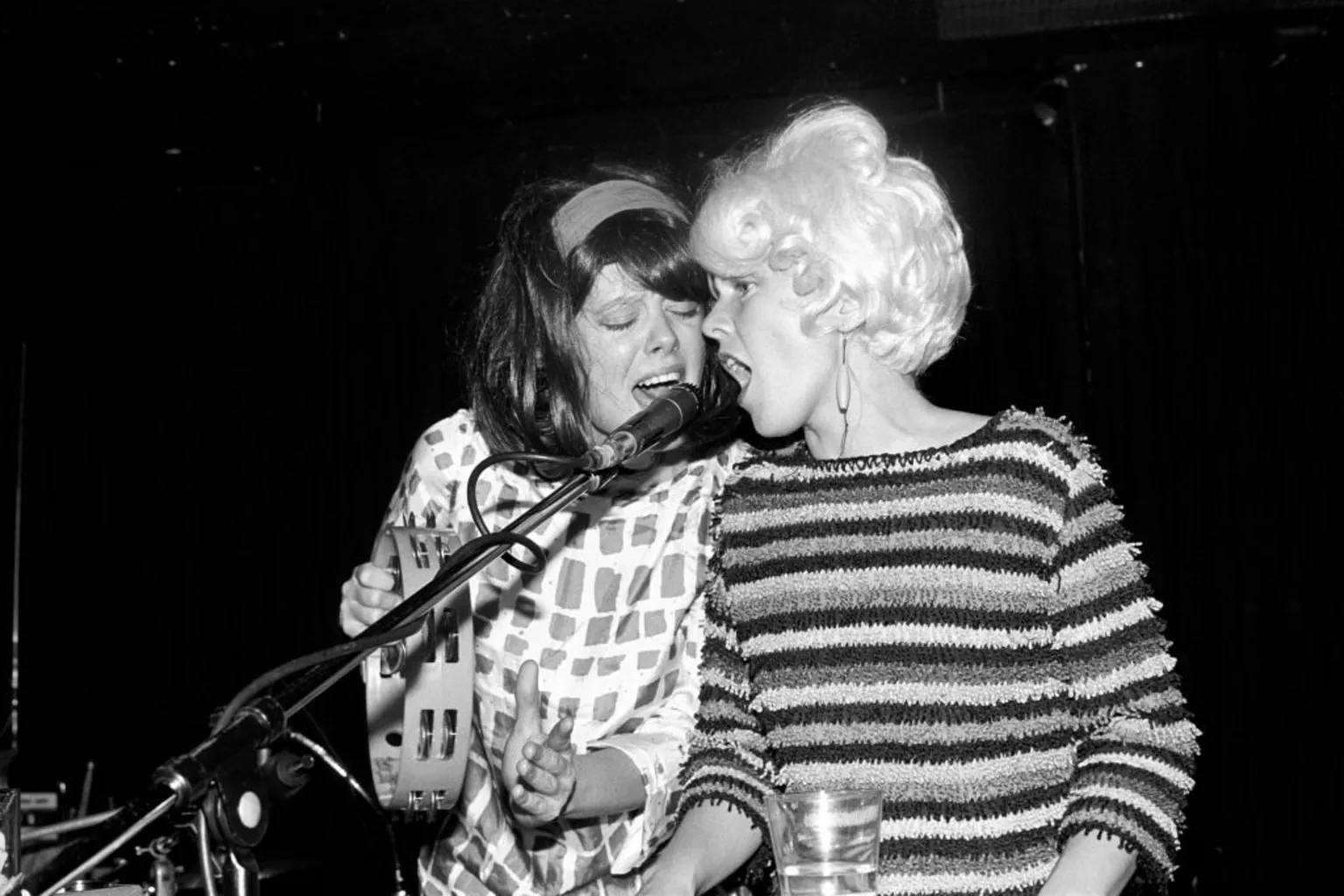  Kate Pierson and Cindy Wilson of The B-52s sing into a single microphone, while wearing retro 1950s outfits and beehive hairdos