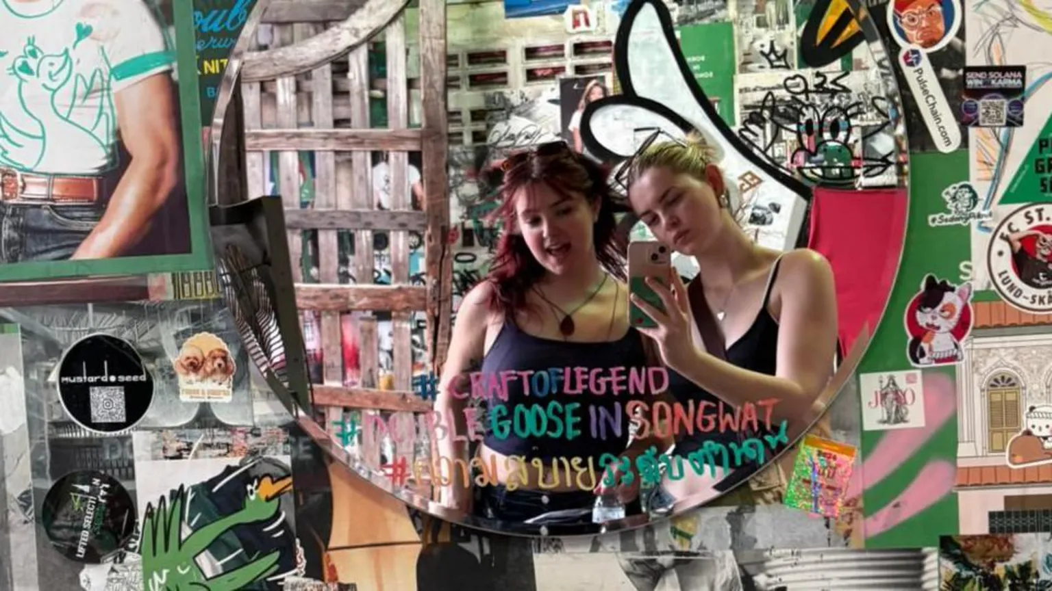 Gwen Shepherd Mia and Gwen taking a selfie of themselves in a mirror. The mirror is on a wall which is covered with various pictures and slogans.