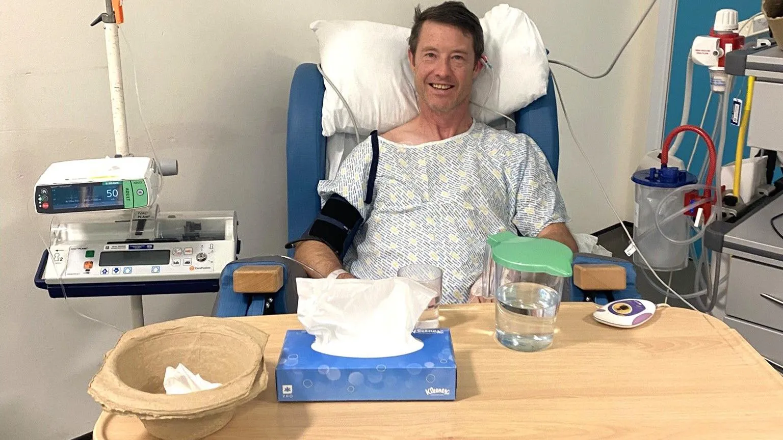 Ian Rodger in hospital, wearing a hospital gown and smiling at the camera.
