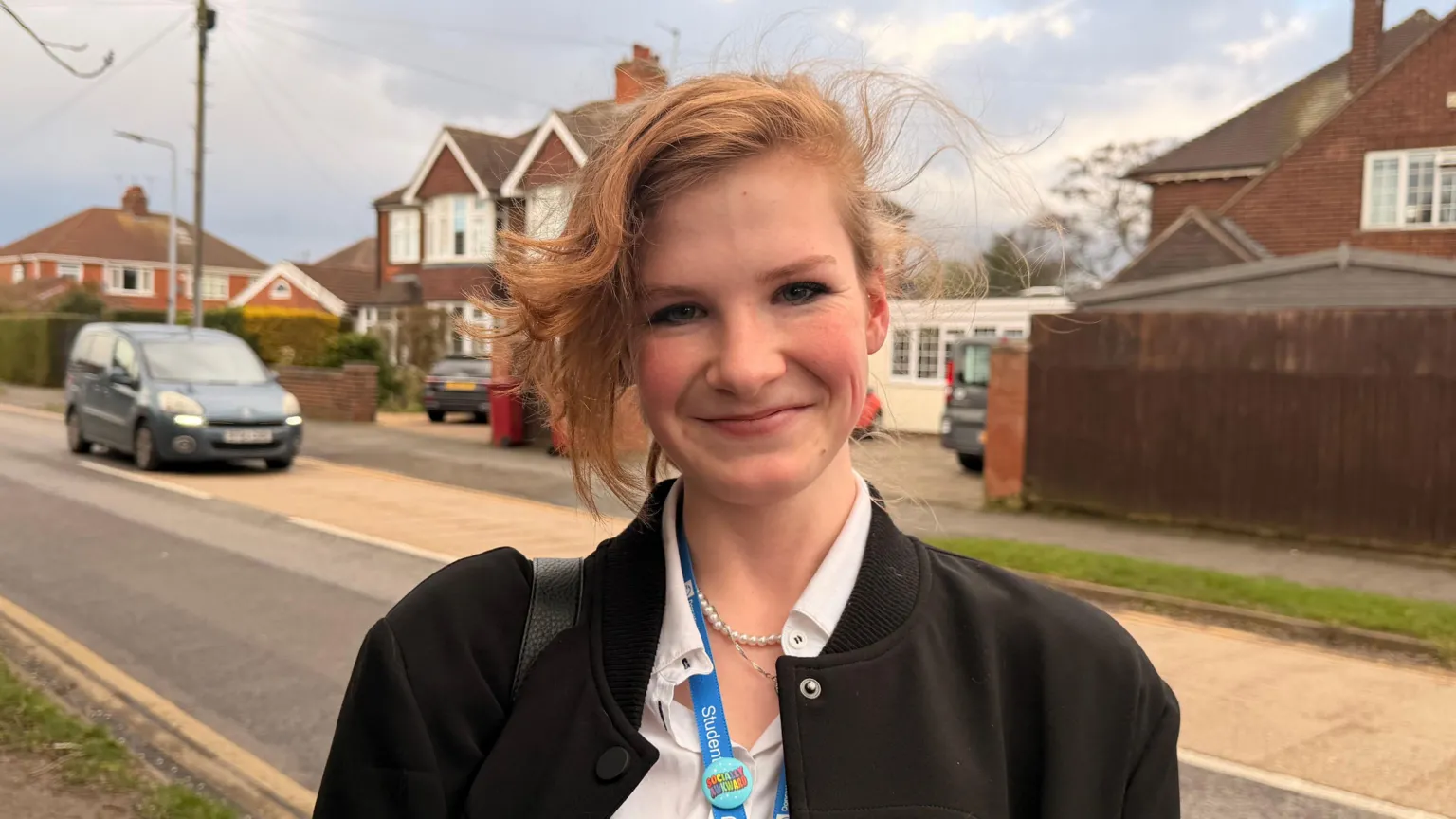 Jasmine Lowe/ A young woman smiles with closed lips as she stands in a residential street with red-brick semi-detached houses behind her. She has ginger hair in an up-do with side parting. She is wearing a black coat with a white shirt, pearl necklace and blue lanyard. 