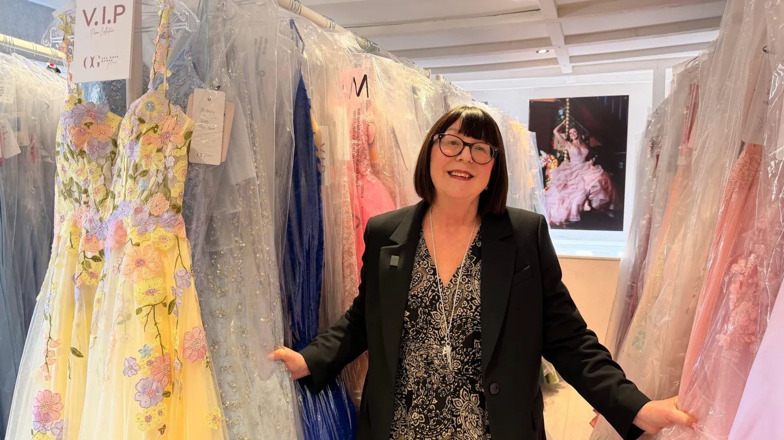Jasmine Lowe/ A woman smiles as she stands between two racks of prom dresses in plastic wrappers, with pink, yellow and blue colours on show. She has a short black bob and a fringe and wears a black and gold floral dress, black blazer and round-rimmed glasses. In the background, a print of a woman on a swing in an elaborate pink gown can be seen.