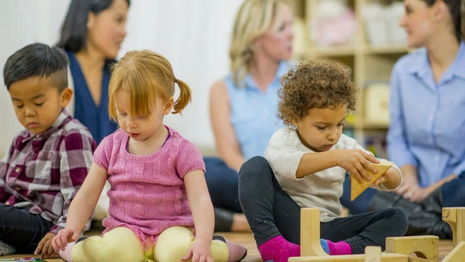  Three young children sit on the floor playing with toys in a library, with their mothers sitting behind.